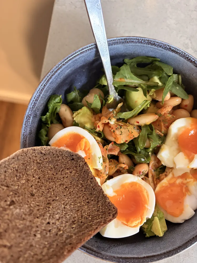 Bowl of butter beans, rocket, avocado, kimchi and jammy eggs with a slice of dark rye